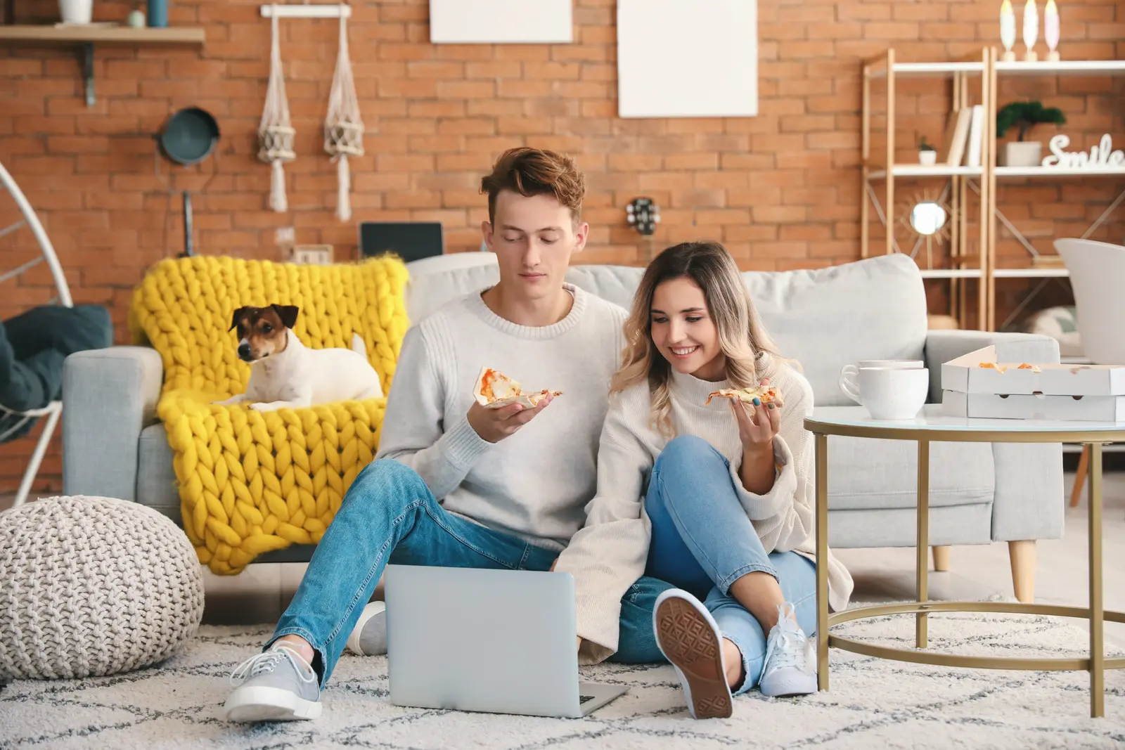 Casal sentado no chão da sala comendo pizza com o notebook ao lado